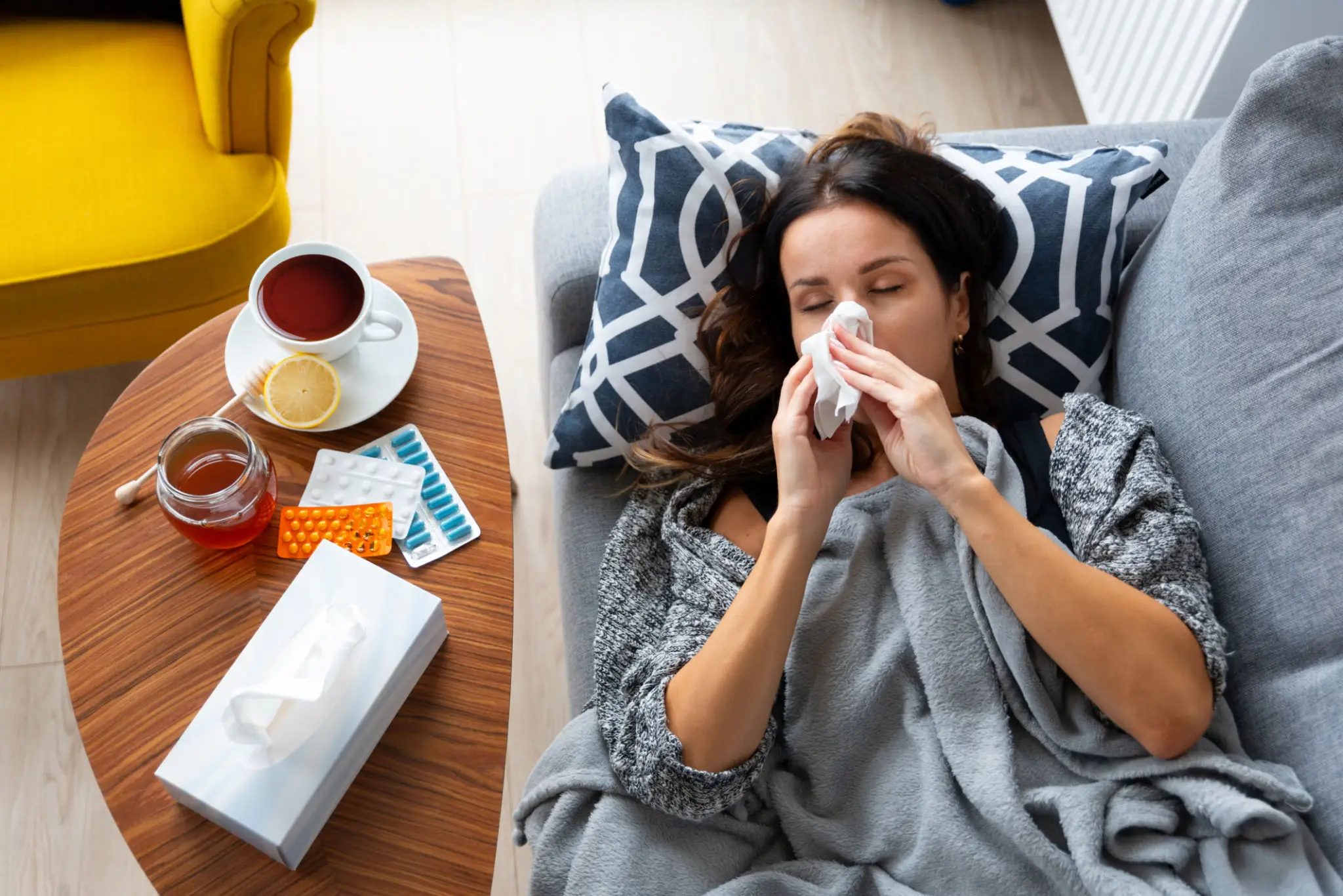 A woman lying in bed, blowing her nose and surrounded by tissues and tea.